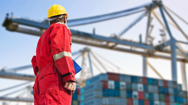 FCL: Mitarbeiter in rotem Overall in Hafen, mit vielen Containern und Lastkränen im Hintergrund FCL: Mitarbeiter in rotem Overall in Hafen, mit vielen Containern und Lastkränen im Hintergrund