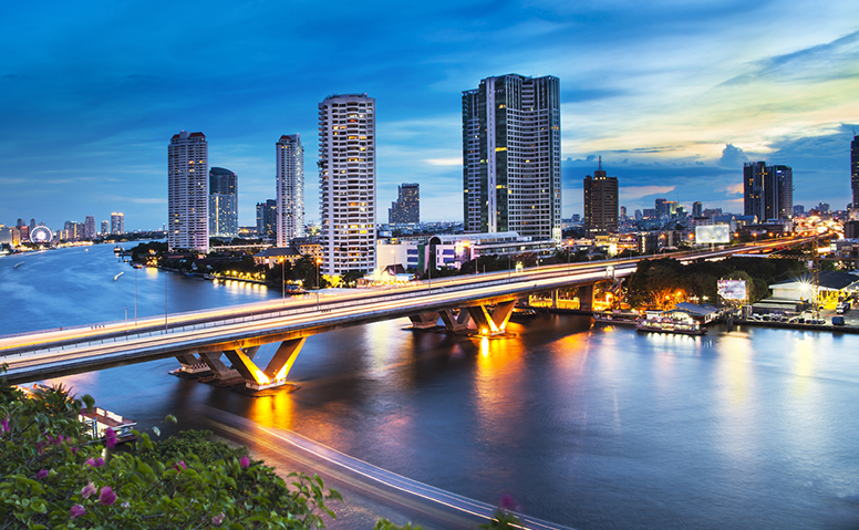 Thailand: Photo of a bridge over the Chao Phraya river in Bangkok, the bridge is illuminated and there are houses and skyscrapers in the background